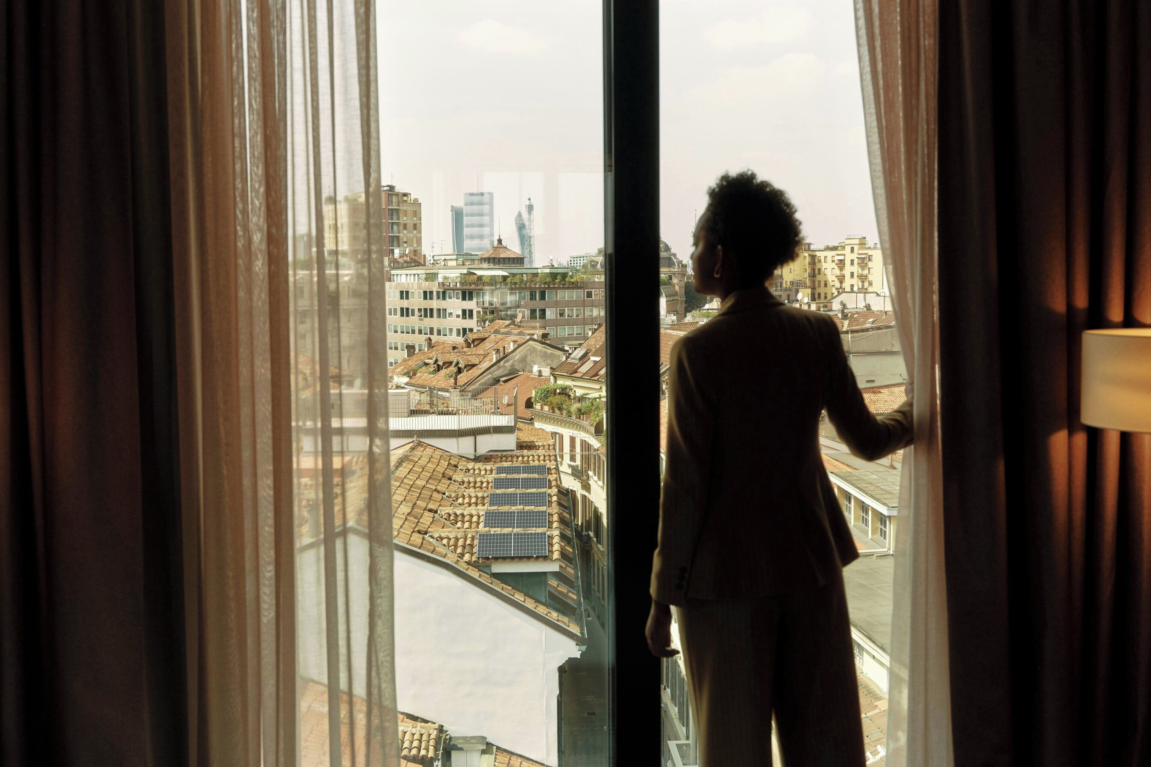 A person standing by a window with a view of a cityscape, including buildings and a bridge.