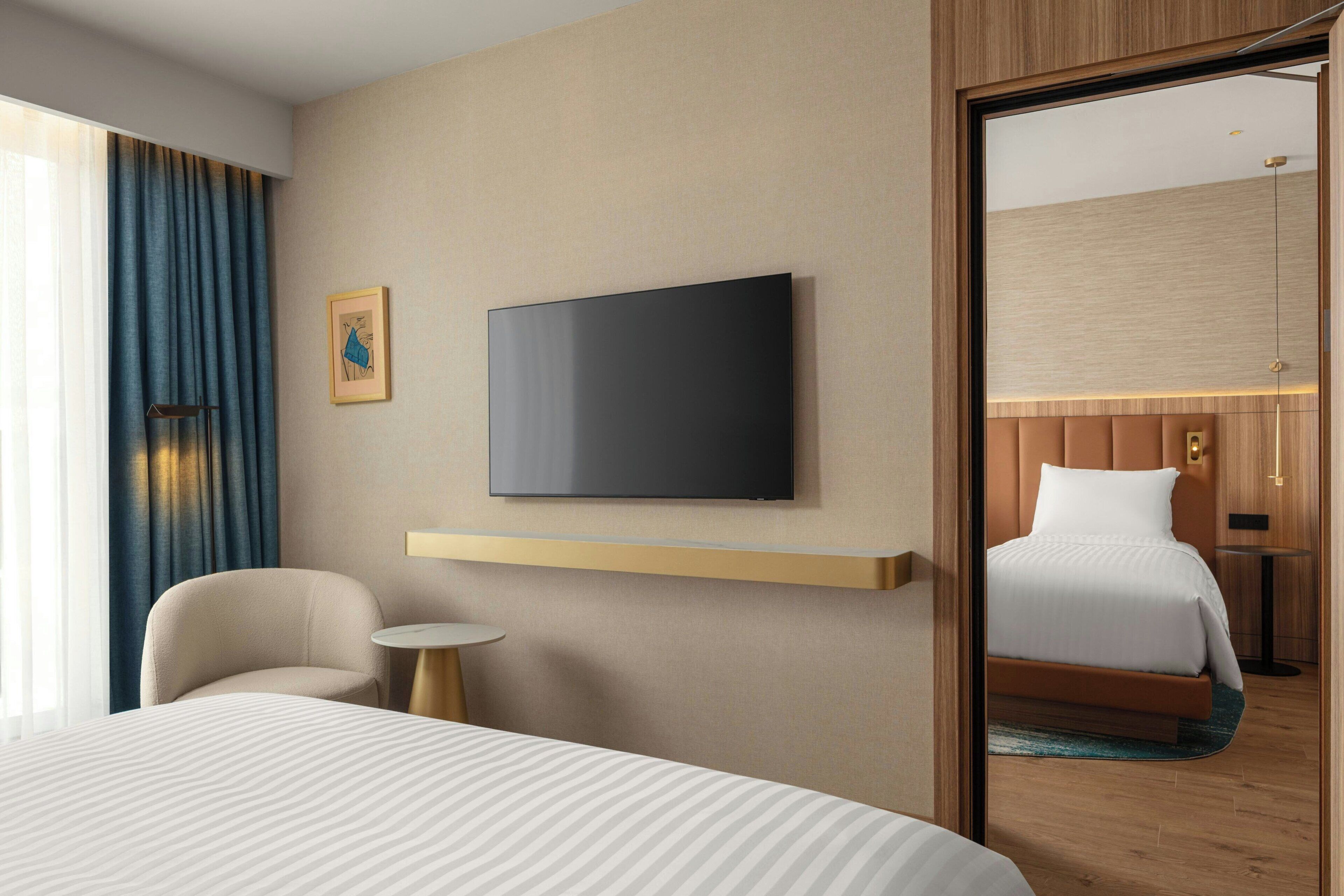 Habitación de hotel con cama, televisor montado en la pared, una mesita redonda, una silla y vista a otra habitación por una puerta abierta.