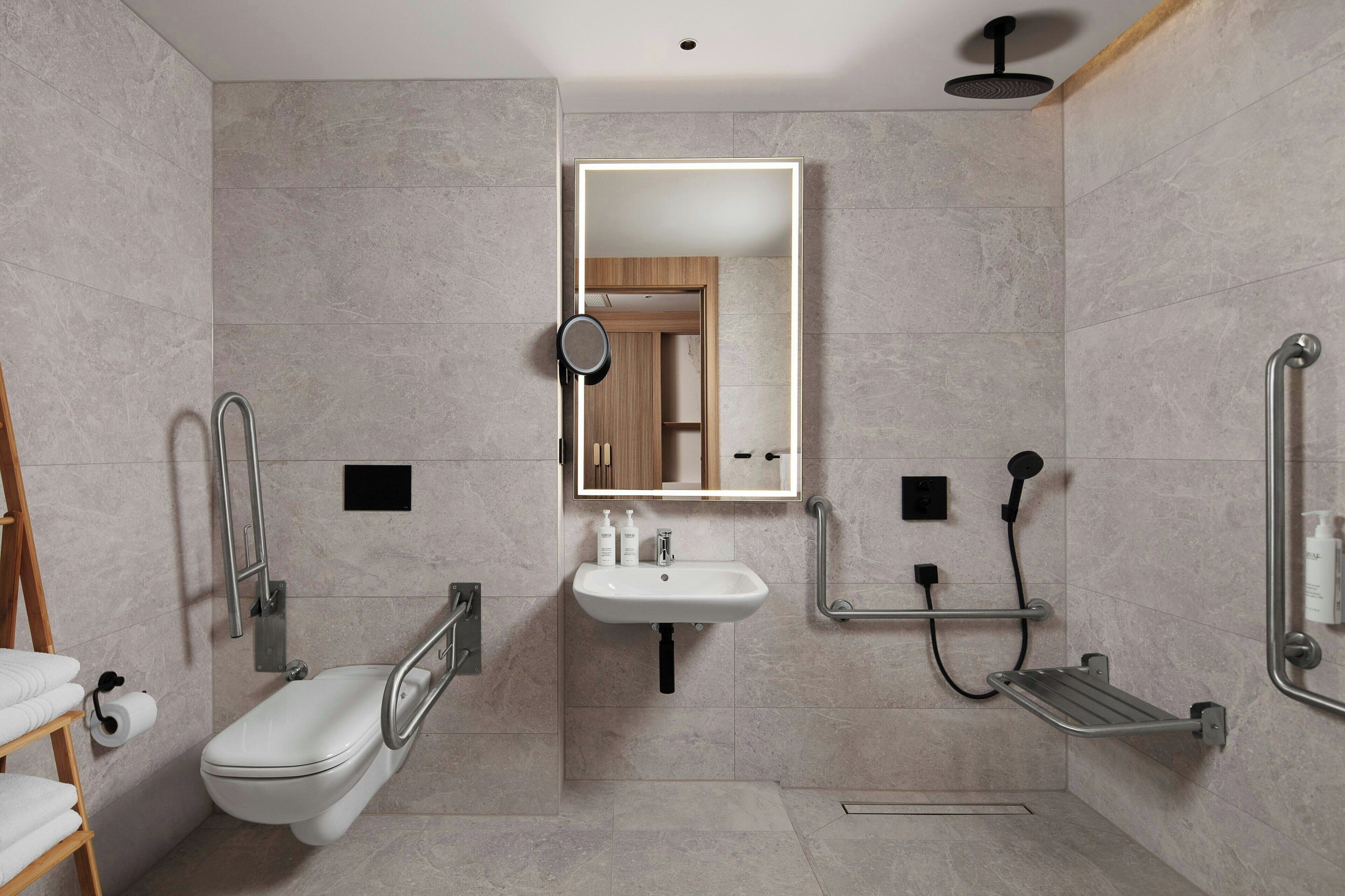A modern bathroom with a toilet, sink, and shower. It includes grab bars and a mirror with lighting.