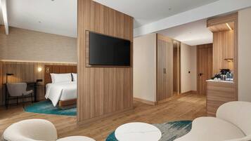 A modern hotel room with a large bed, a TV mounted on a wooden panel, a desk with a chair, and a small round table.