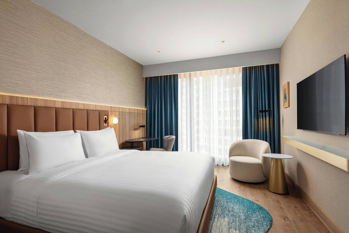 Ein Hotelzimmer mit einem großen Bett, einem Schreibtisch, einem Sessel, einem Fernseher und einem Fenster mit Vorhängen.