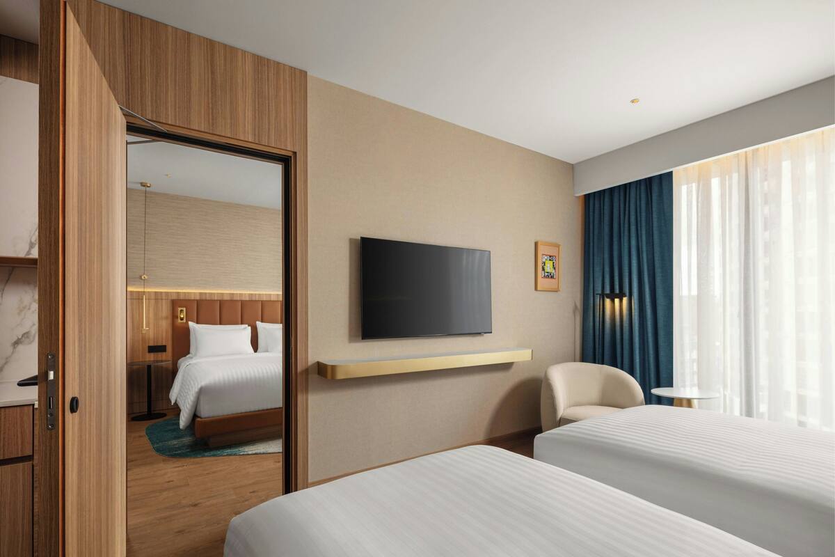 Ein Hotelzimmer mit zwei Betten, einem Fernseher, einem Schreibtisch, einem Sessel und einem großen Fenster mit Vorhängen.
