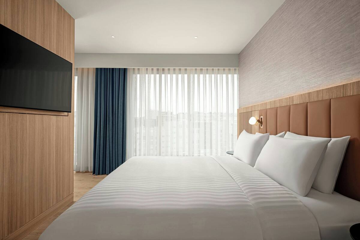 Ein modernes Hotelzimmer mit einem großen Bett, einem flachbildfernseher, der an einer Holzplatte befestigt ist, Blick auf die Stadt durch transparente Vorhänge und einer Wandleuchte.