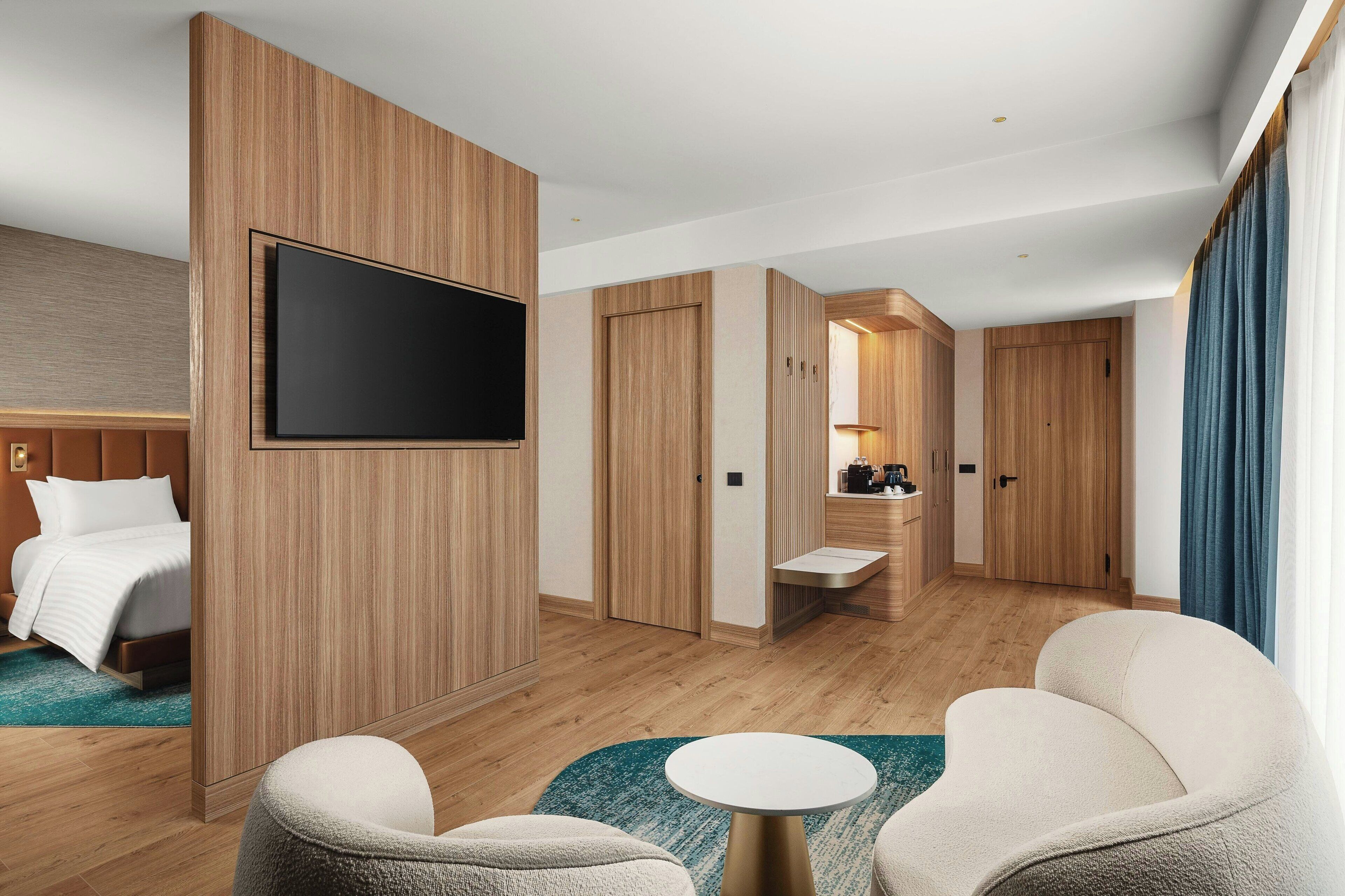 A modern hotel room with a large bed, a flat-screen TV mounted on a wooden panel, a seating area with a round table, and a kitchenette in the background.