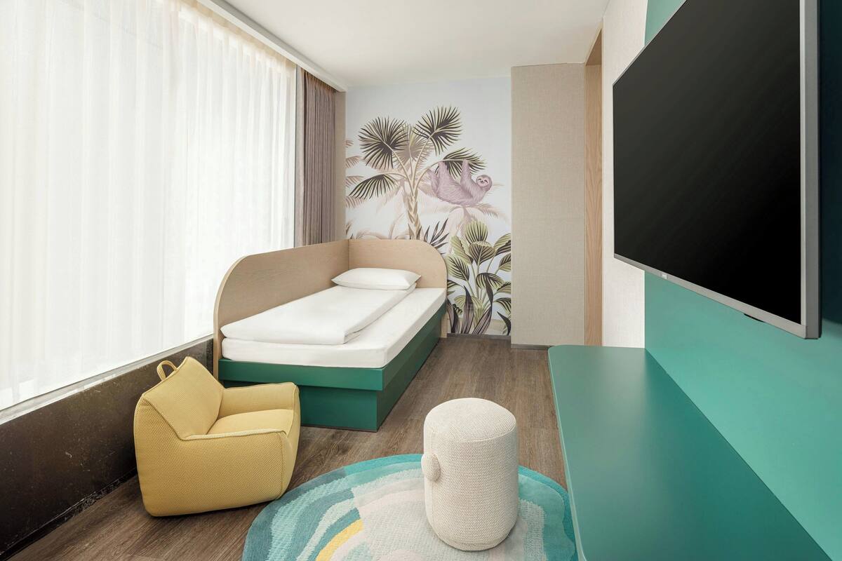 A modern bedroom with a bed, a yellow armchair, a round ottoman, and a large TV.