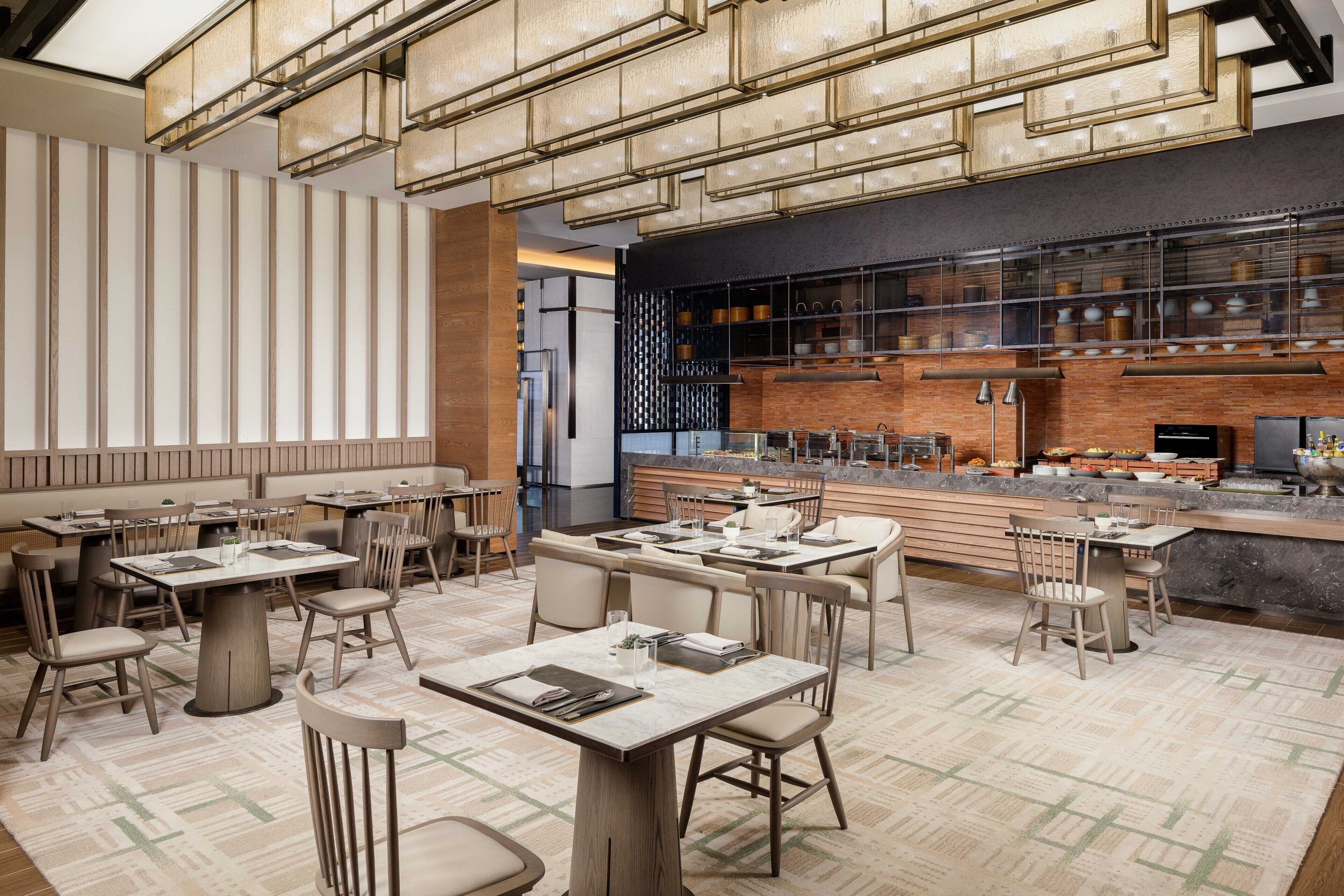 A modern restaurant with multiple dining tables and chairs, a kitchen area with visible cooking equipment, and a patterned floor.
