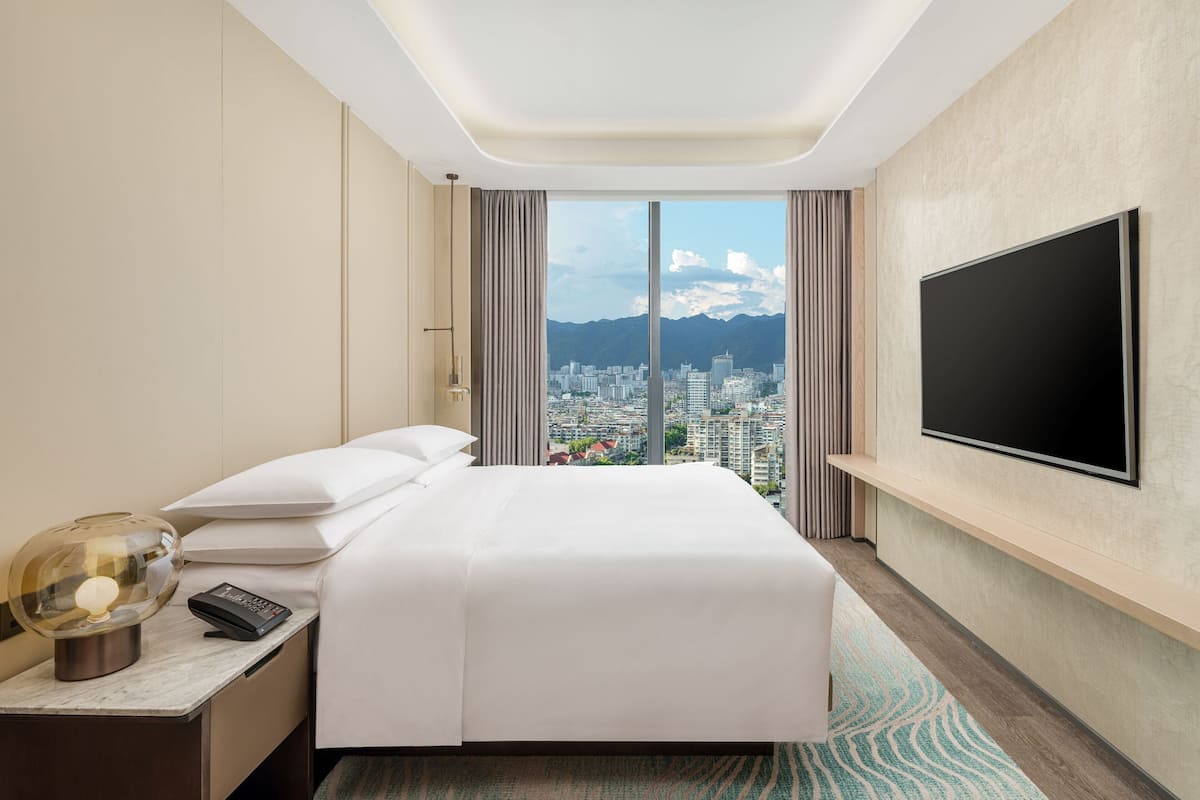 A hotel room with a large bed, a TV mounted on the wall, a nightstand with a lamp, and a view of a cityscape through a large window.