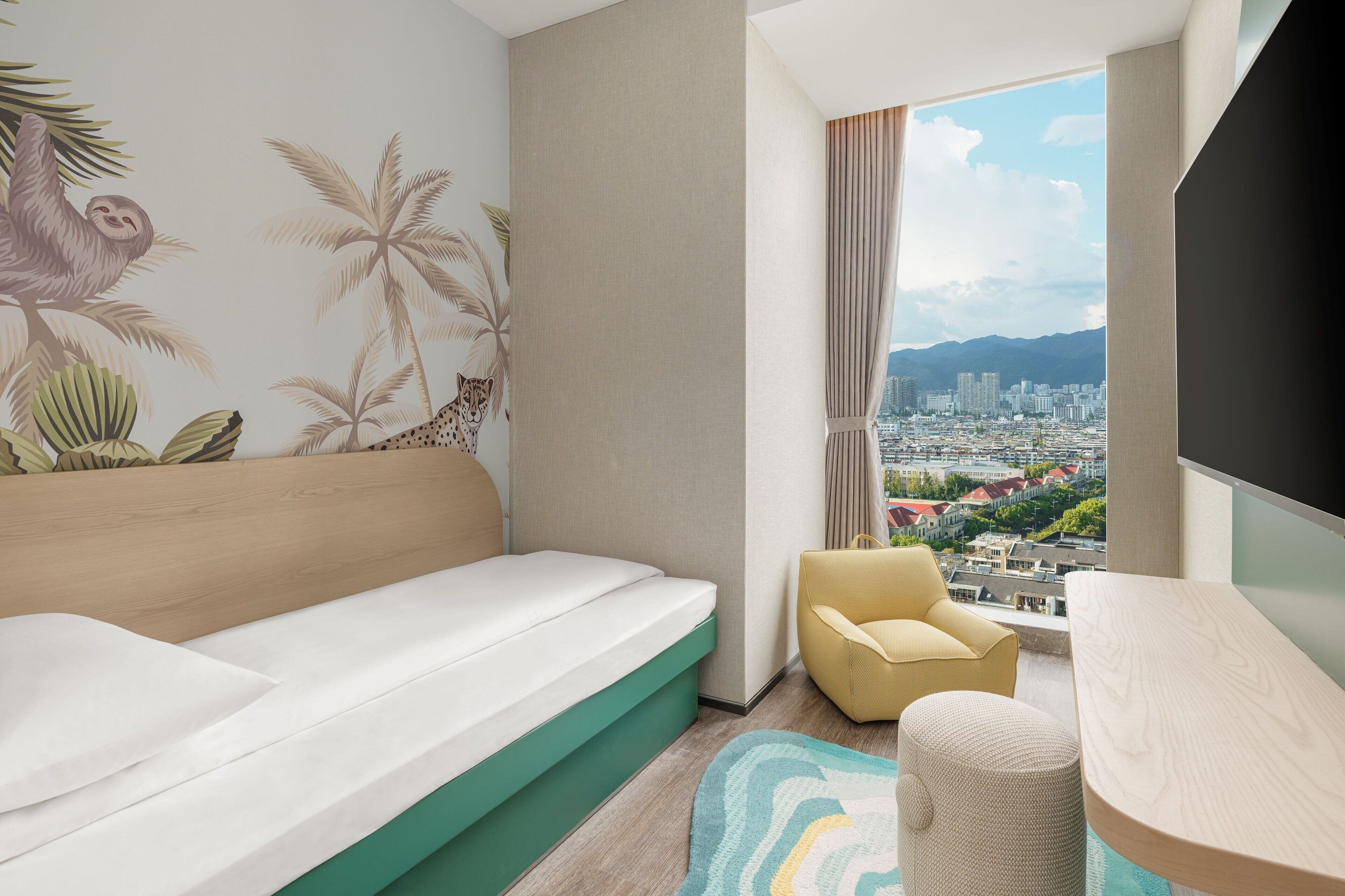 A modern hotel room with a bed, a chair, a small table, a view of a cityscape, and a wall with tropical mural art.