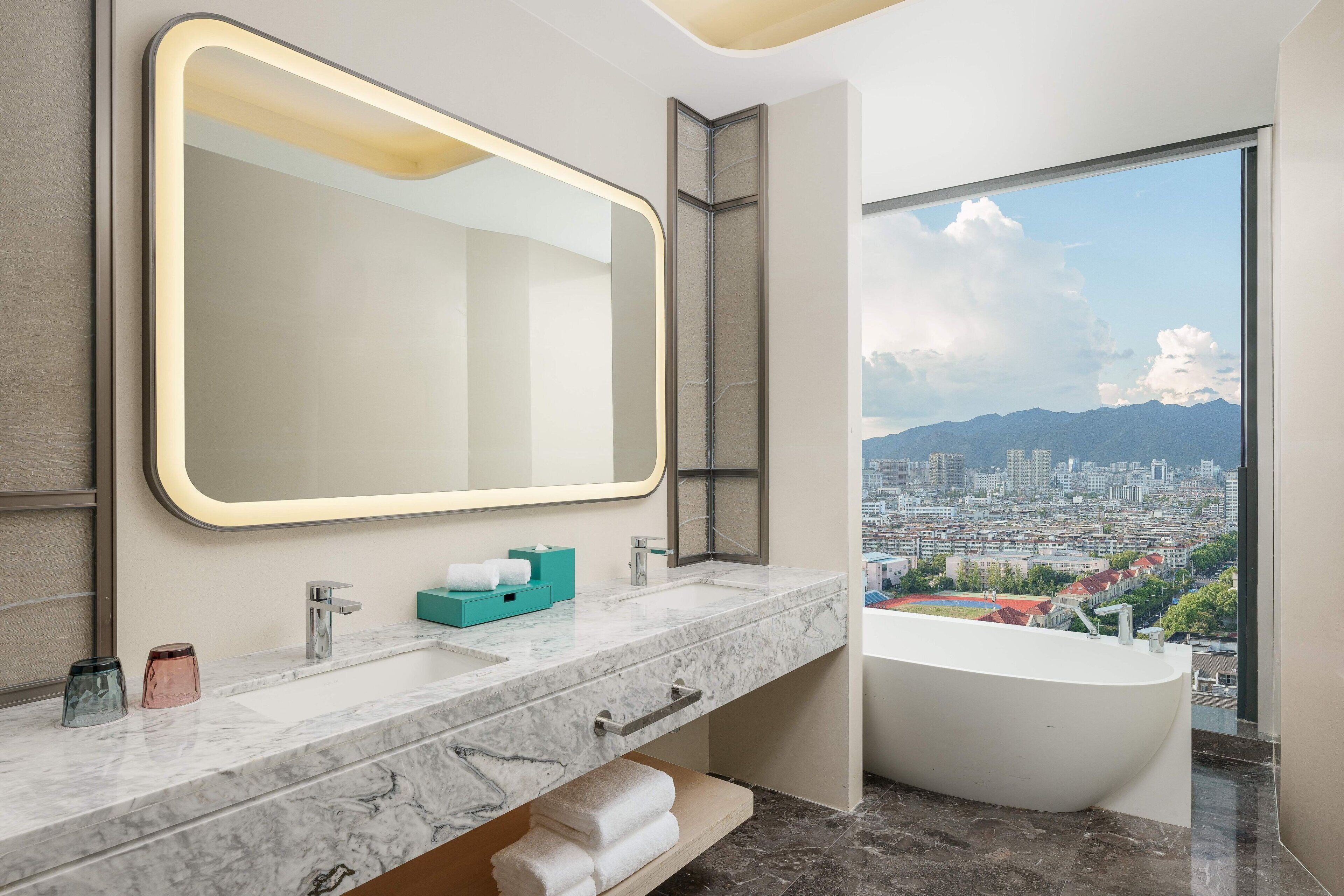 Um banheiro moderno com um espelho grande, uma banheira e vista para a cidade.