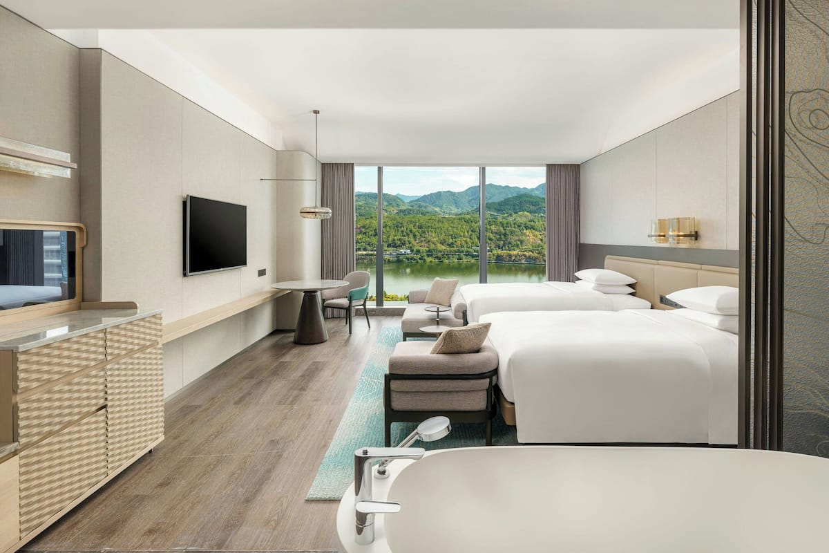 A modern hotel room with a large bed, a separate sofa, a flat-screen TV, a desk, and a view of a lake and mountains.