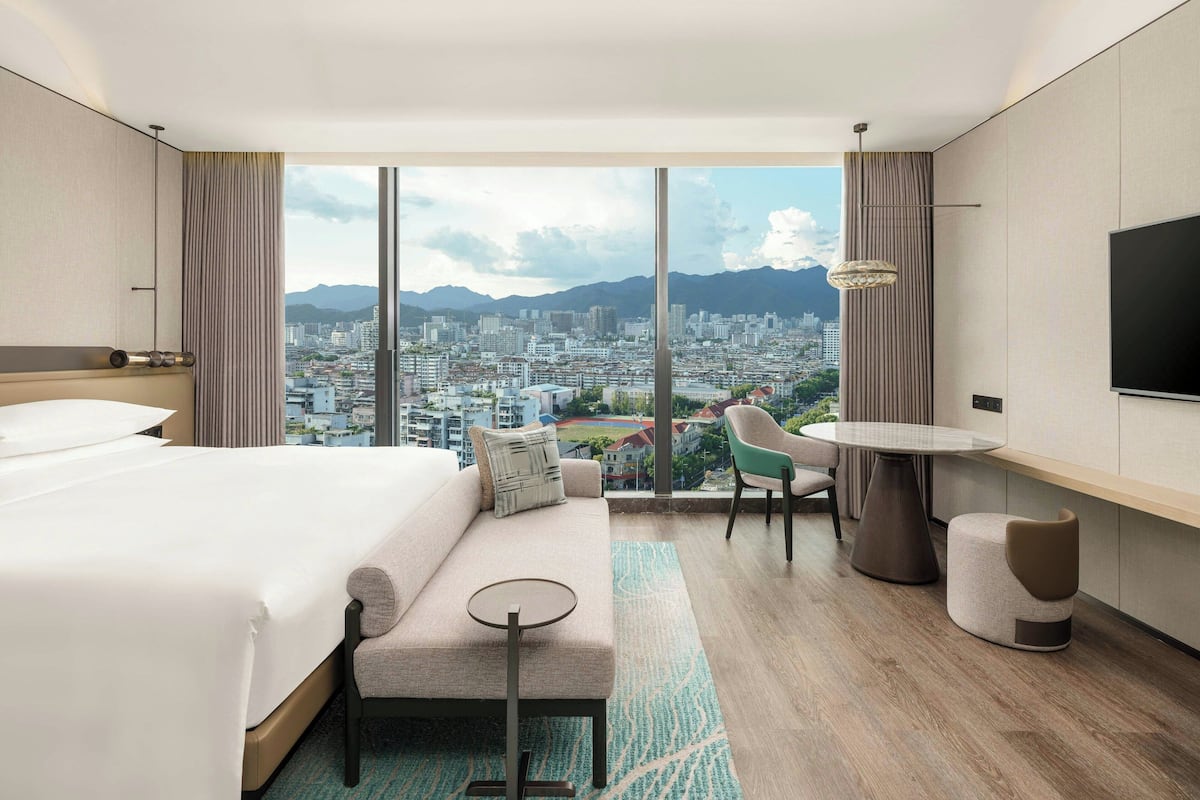 A modern hotel room with a large bed, a sofa, a small round table, and a chair. There is a large window offering a city view.