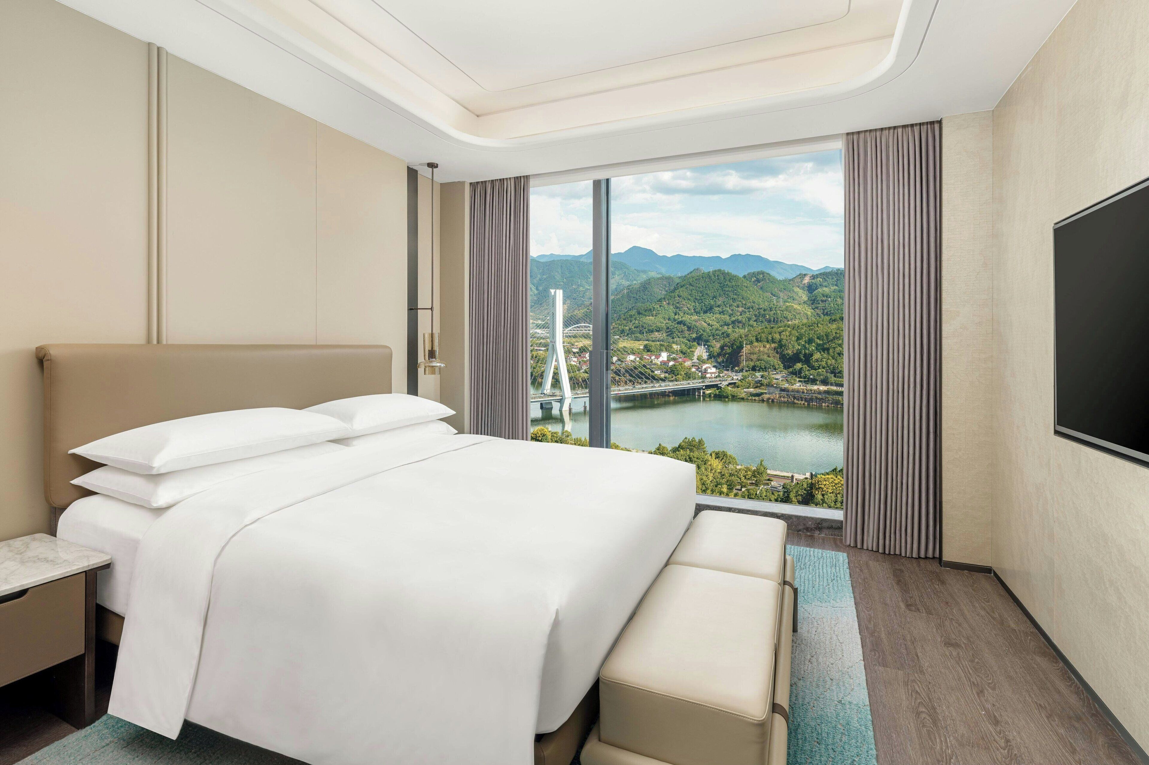 A hotel room with a large bed, a TV, and a view of a bridge and mountains.