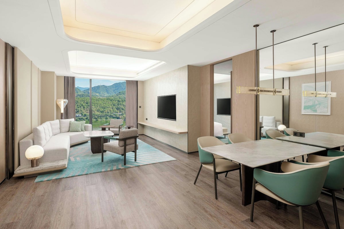 A modern hotel room with a sofa, dining table, chairs, a flat-screen TV, and a large window offering a mountain view.