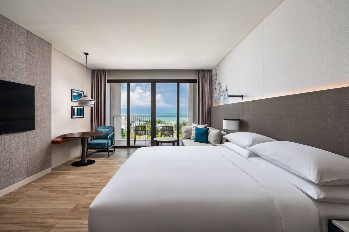 Una habitación de hotel moderna con una cama grande, una zona de estar con una mesa y sillas, un televisor montado en la pared y un balcón con vista al océano.