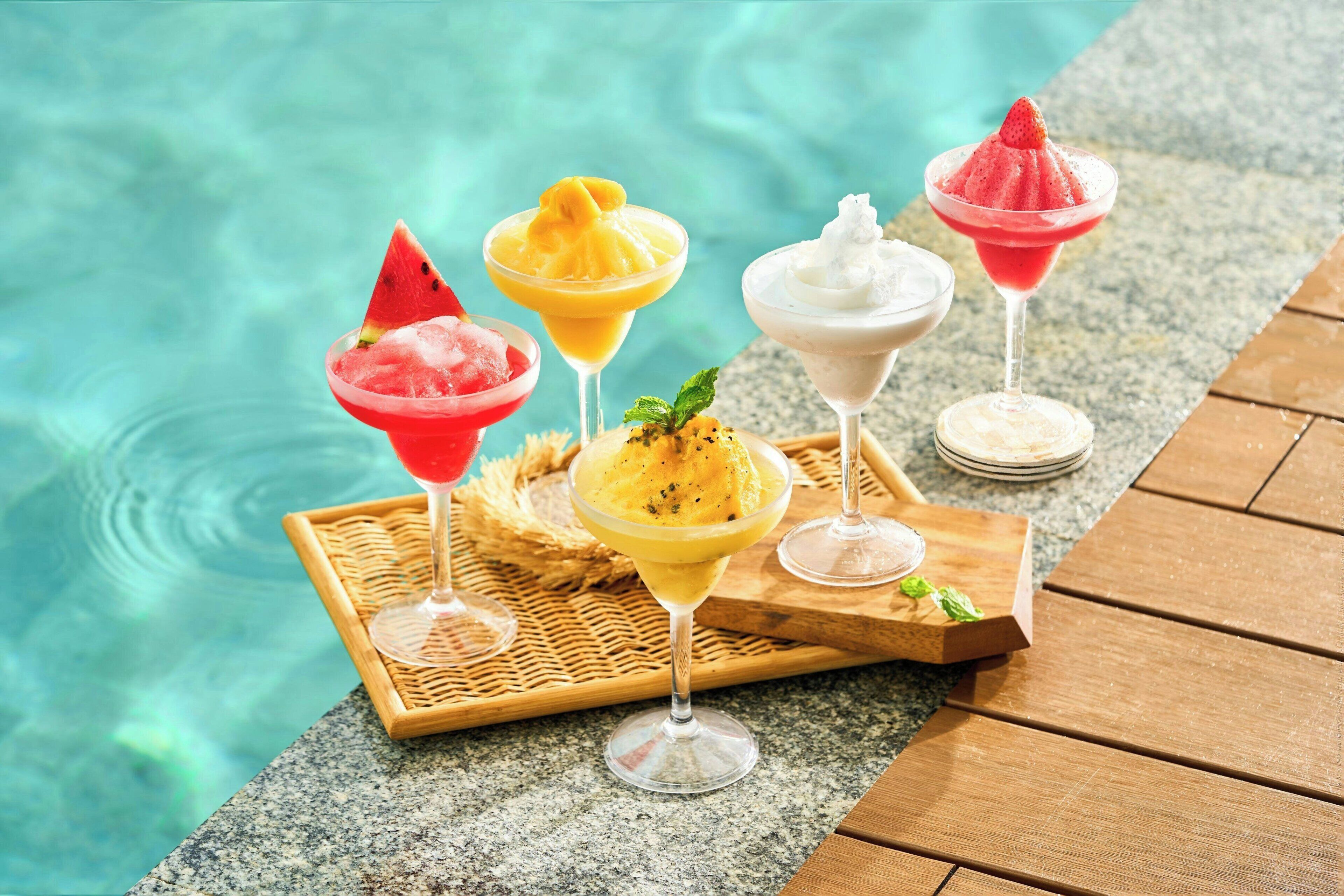 A poolside setting with various fruit-based cocktails in glasses, a wicker tray, and a wooden table.