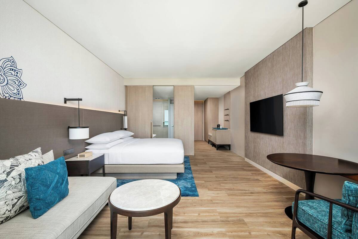 Una habitación de hotel moderna con una cama, un sofá, una mesita, un televisor montado en la pared y un pasillo que conduce a un baño.