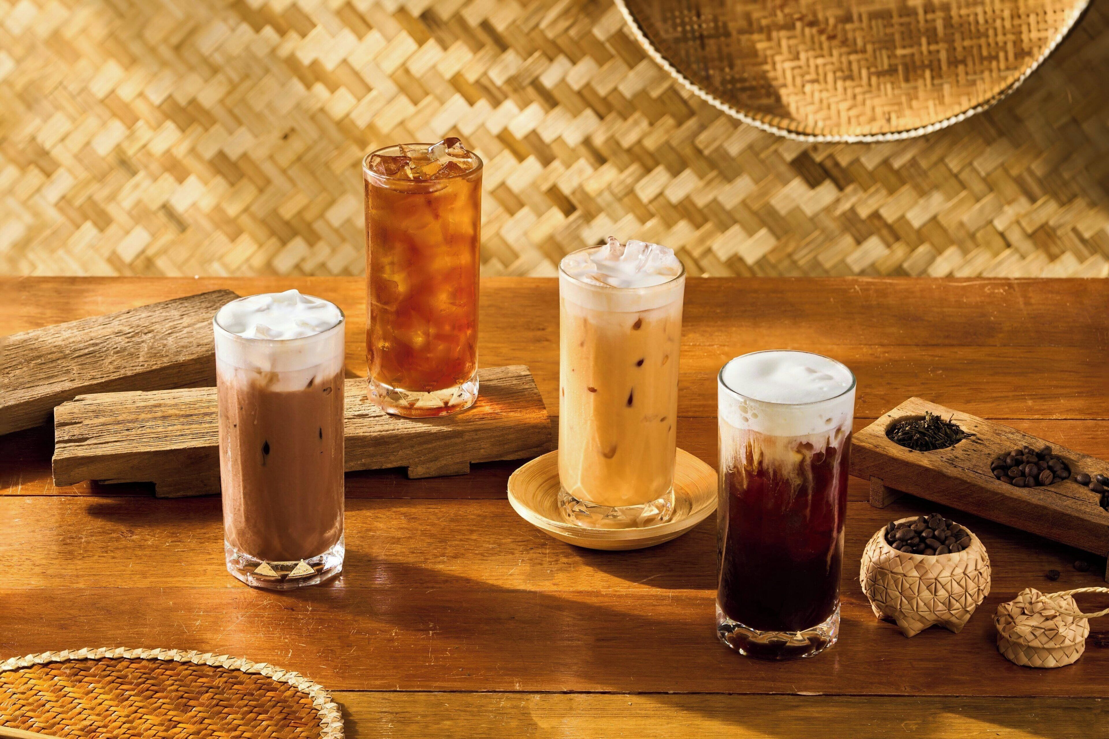 Cuatro bebidas heladas en vasos sobre una mesa de madera con esteras tejidas y granos de café al fondo.