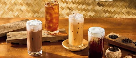 Cuatro bebidas heladas en vasos sobre una mesa de madera con esteras tejidas y granos de café al fondo.