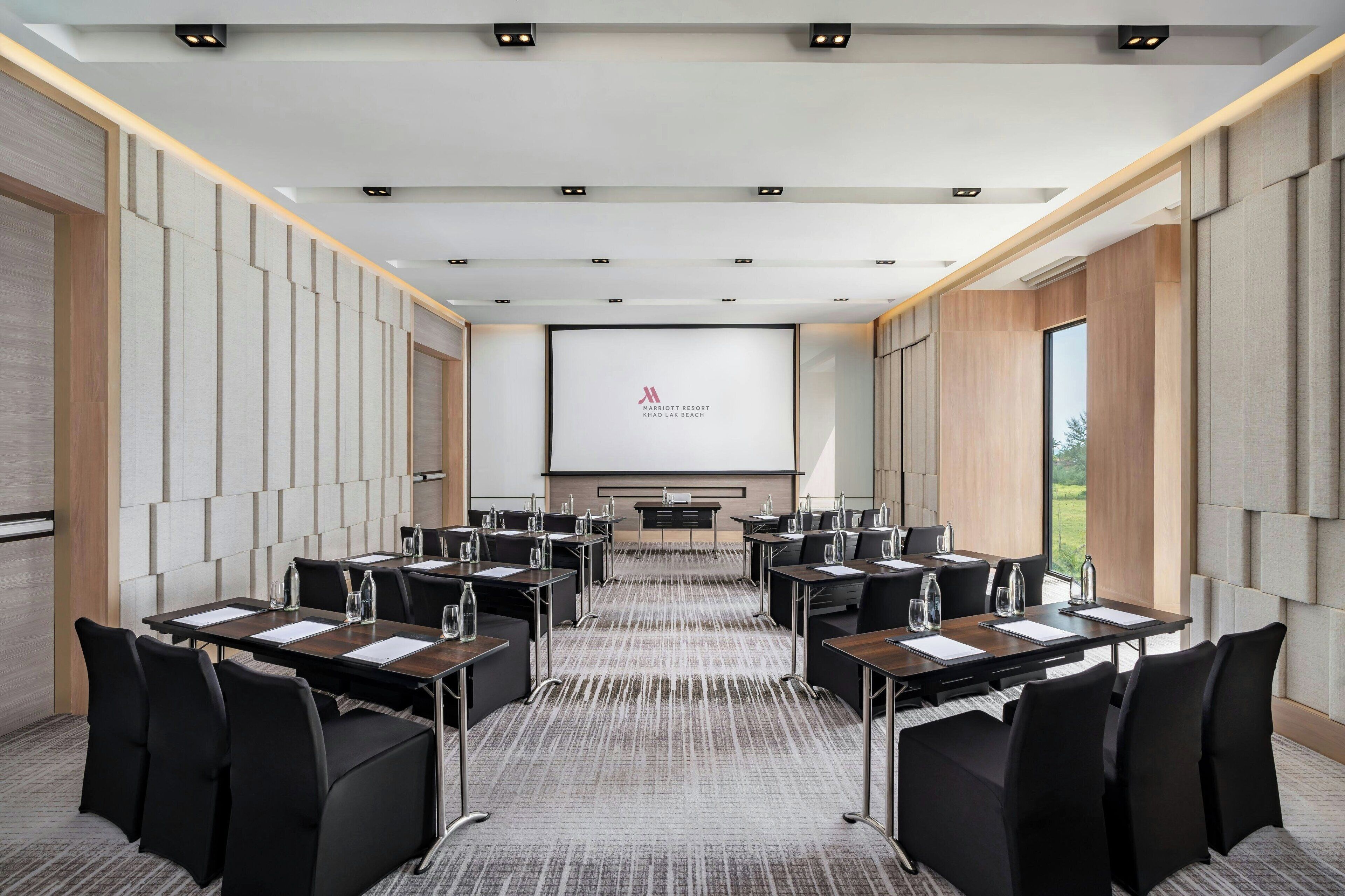 A conference room with a large screen, multiple tables, and chairs, featuring a neutral color scheme and modern design.