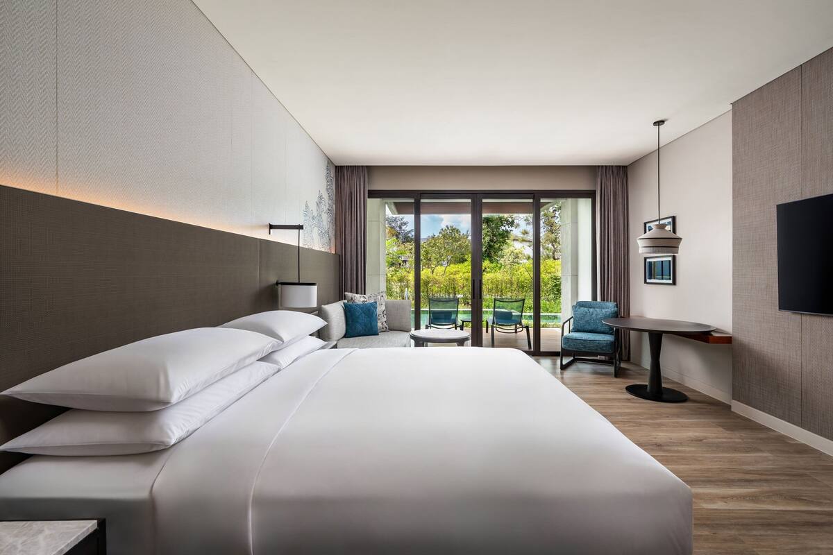 Habitación de hotel moderna con una cama grande, una mesita, dos sillas, un televisor y un balcón con vistas a zonas verdes.