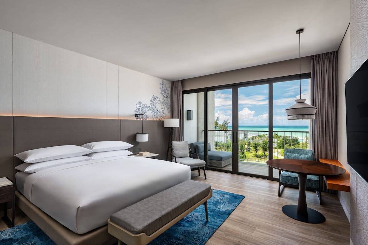 Una habitación de hotel moderna con una cama grande, un balcón con vista al océano, una zona de estar con una mesa y un televisor.