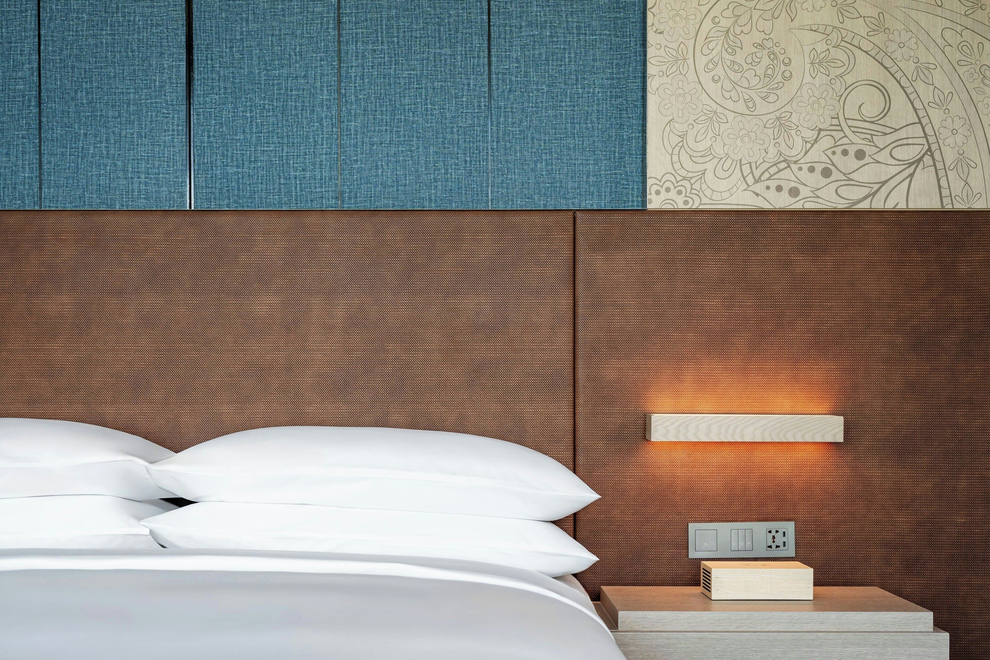 A neatly made bed with white linens, a wooden nightstand with a power outlet, and a wall-mounted light fixture.
