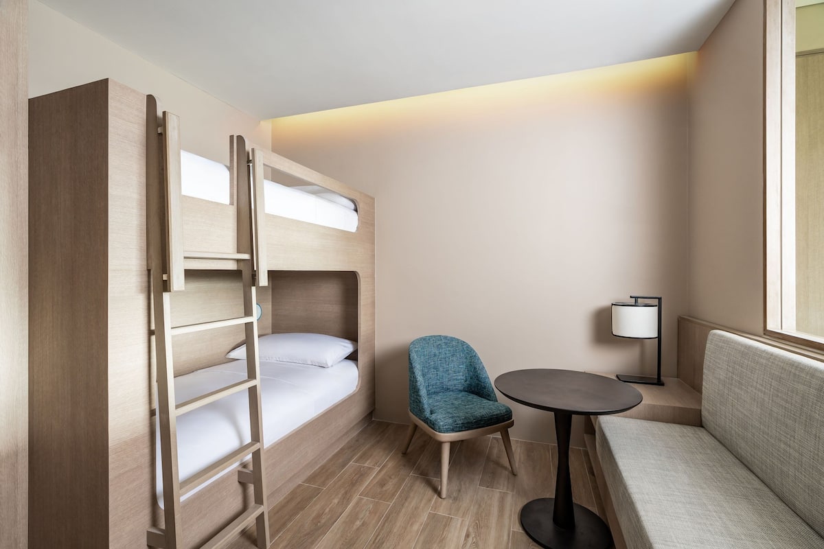 Una habitación de hotel pequeña con una litera, una mesita redonda, una silla y una lámpara de noche.