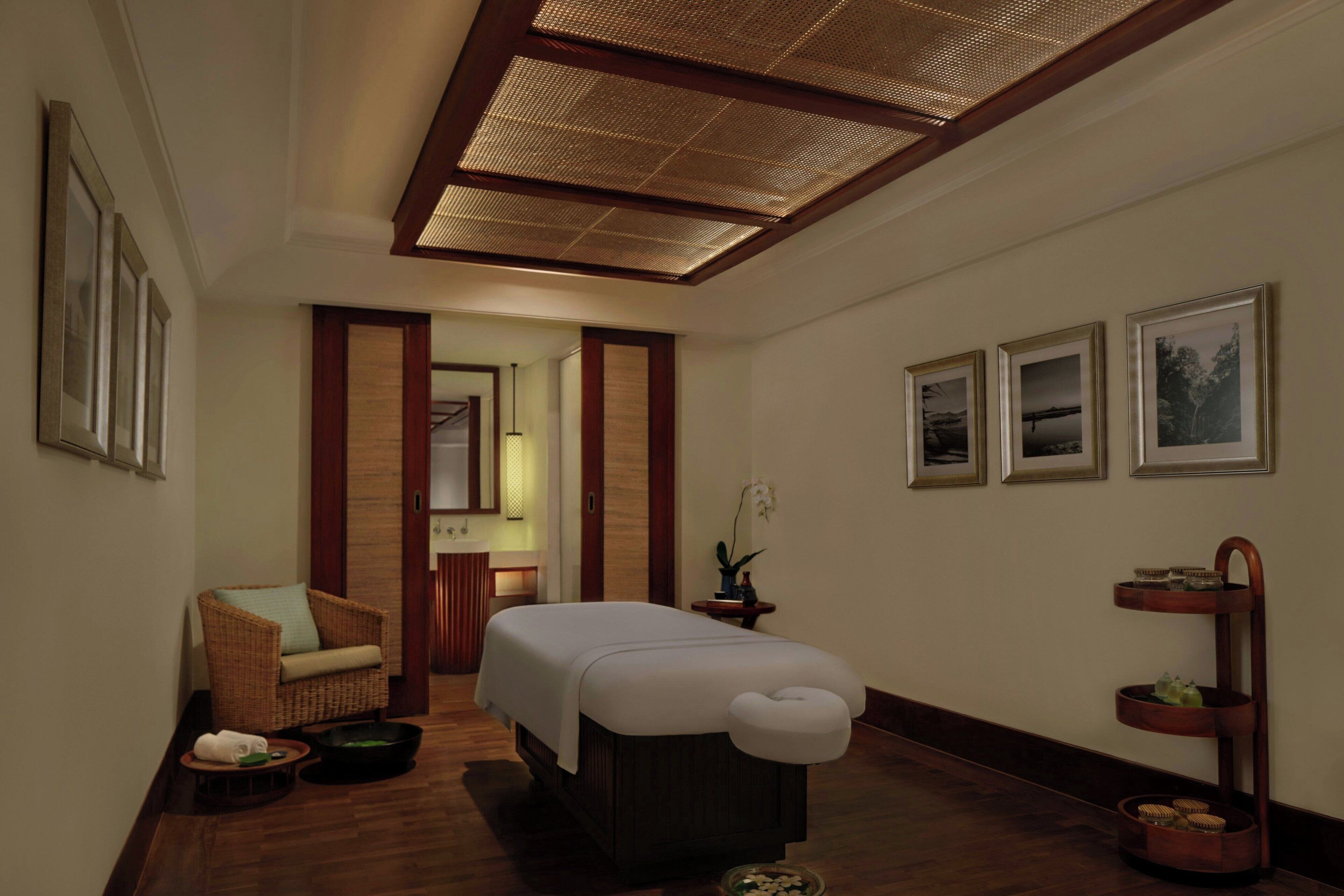 A hotel room with a bed, a chair, a small table, and a shelf with decorative items.