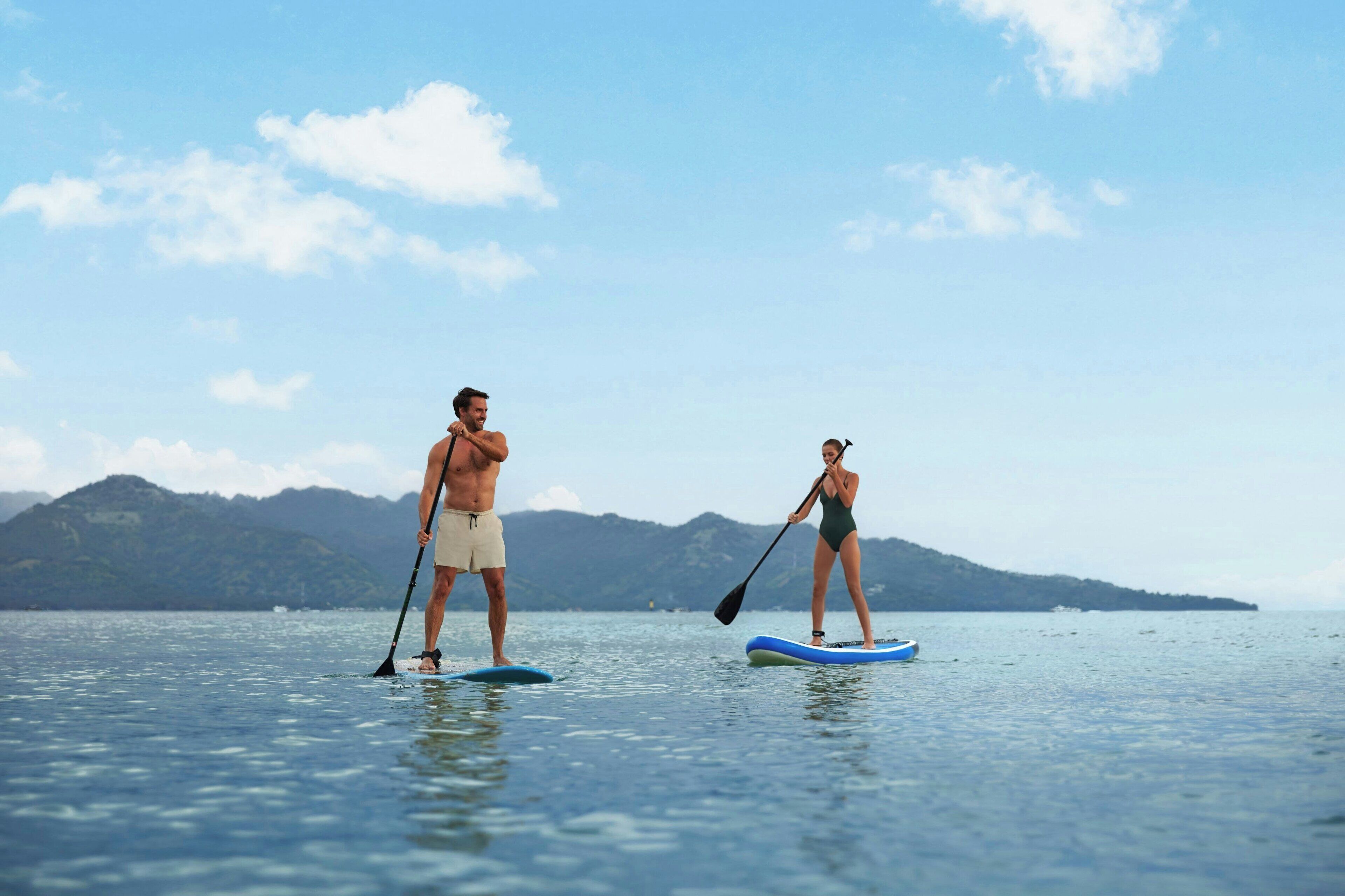 Deux personnes pratiquent le stand-up paddle sur une étendue d’eau calme, avec des montagnes en arrière-plan.