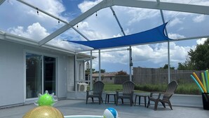 A covered outdoor swimming pool with a blue shade sail, surrounded by patio furniture and chairs. - Family Getaway - Private Pool - Sleeps 8 (New Port Richey)