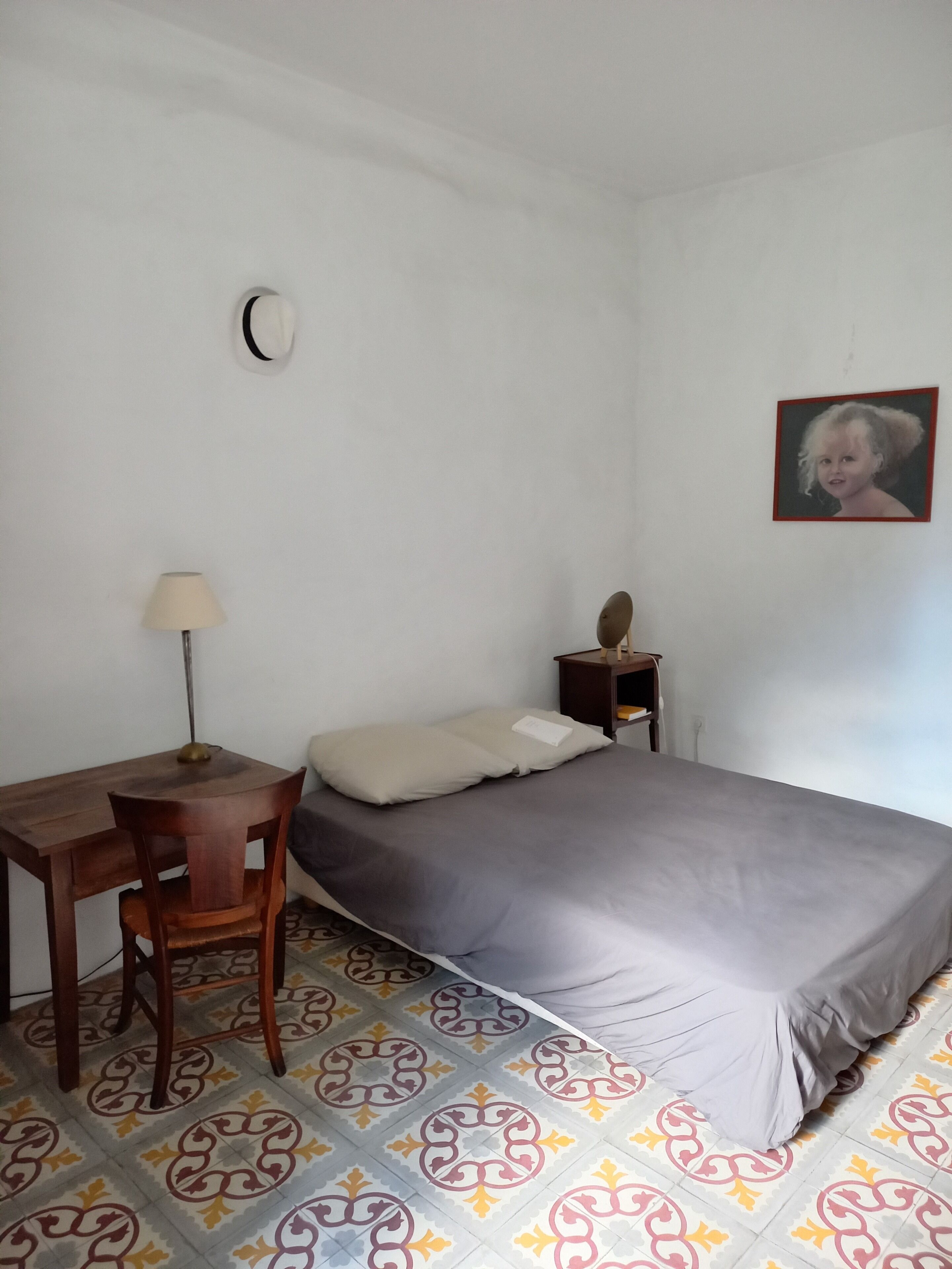 A bedroom with a bed, a desk, a chair, a lamp, a nightstand, and a wall-mounted picture.