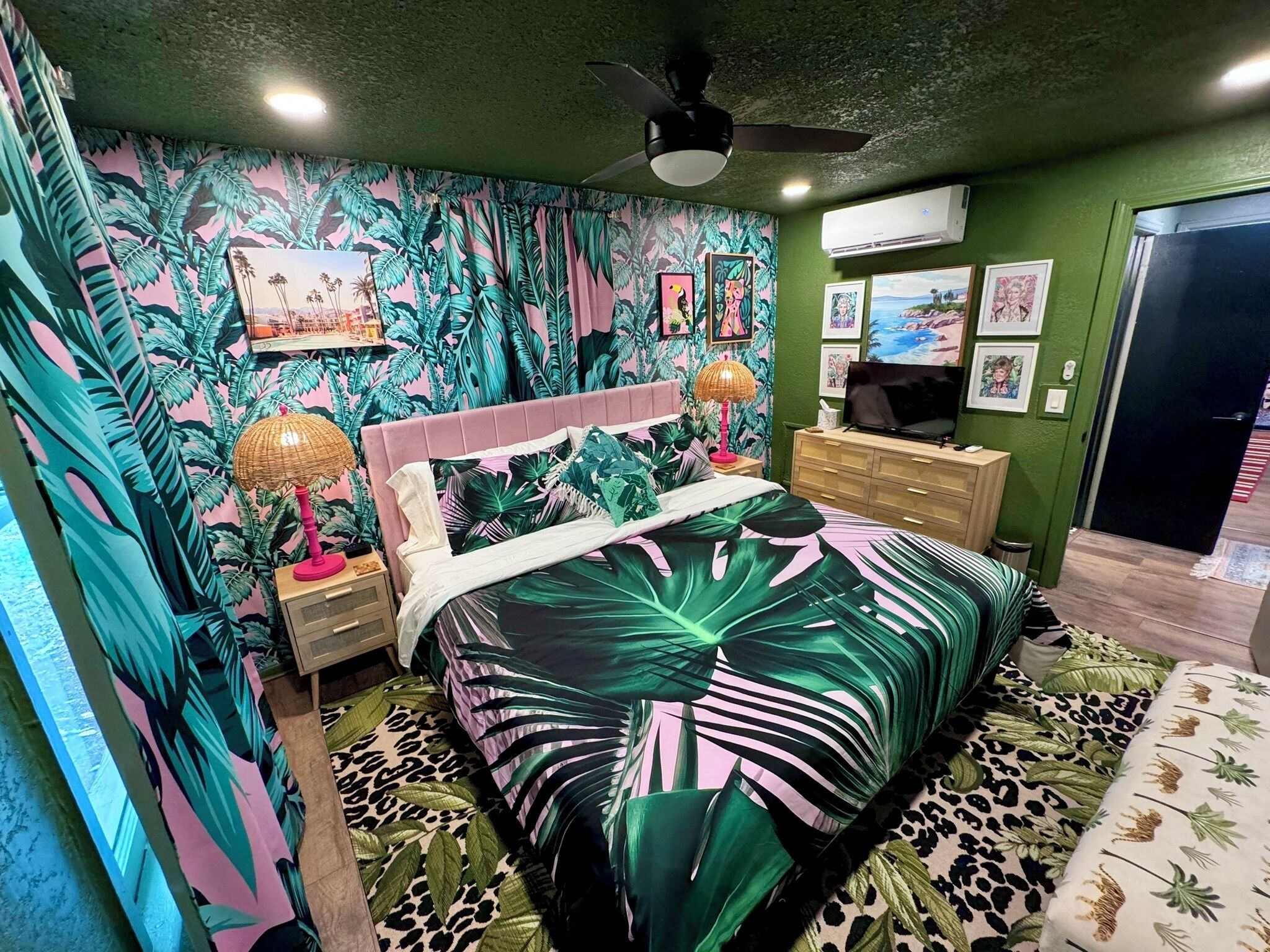 A bedroom with a bed, a nightstand, a dresser, a ceiling fan, and a wall with tropical wallpaper.