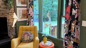 A cozy living room with a yellow armchair, a small round table with a vase, and a window with floral curtains. - Welcome to The Kitschy Kottage (Rogersville)