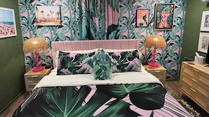 A bedroom with a bed, two nightstands, a dresser, and a wall with tropical wallpaper and framed pictures. - Welcome to The Kitschy Kottage (Rogersville)