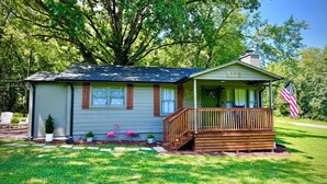 A small, single-story house with a porch, a mailbox, and a small garden with flamingos. - Welcome to The Kitschy Kottage (Rogersville)
