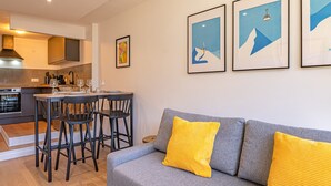A modern living room with a grey sofa and yellow cushions, a dining area with a table and chairs, and a kitchen with a stove and sink.