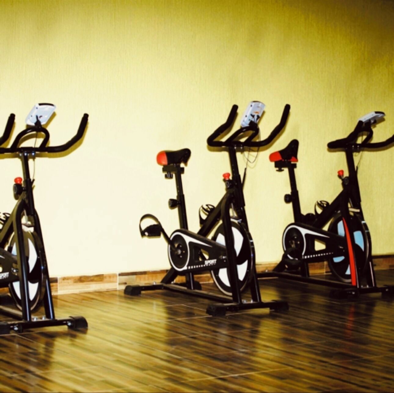 Three stationary bikes in a room with a wooden floor and a plain yellow wall.