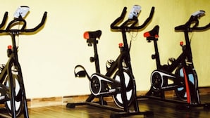 Trois vélos d’appartement sont disposés dans une pièce dont le sol est en bois et un des murs est peint en jaune uni.