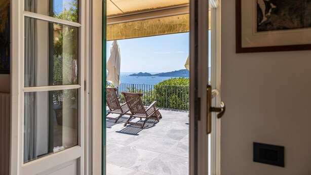 An open door leading to a patio with a view of the sea, outdoor furniture, and a framed artwork on the wall.