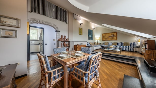 Een eetgedeelte met een houten tafel en rieten stoelen, een woonkamer met een bank en fauteuils, en een keuken die door een boog zichtbaar is.