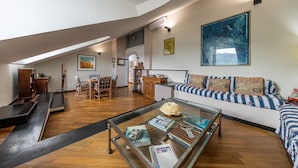 Een ruime loft met hoge plafonds, een groot raam, een bank met gestreepte kussens, een glazen salontafel met tijdschriften, en een eethoek met een houten tafel en stoelen.