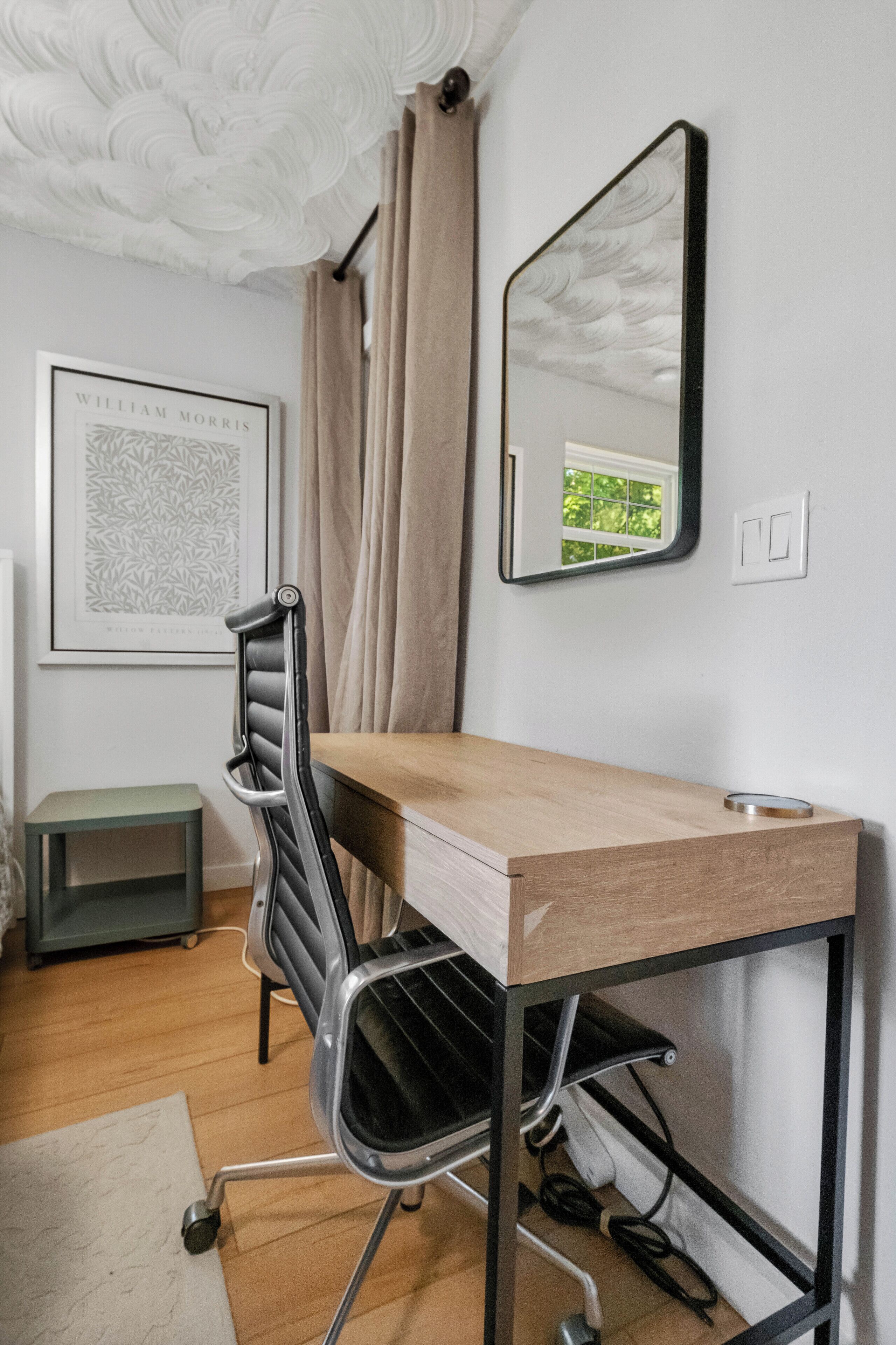 A modern office space with a wooden desk, a black office chair, a framed picture on the wall, and a window with curtains.