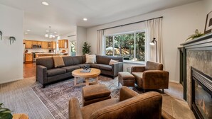 A modern living room with a sofa set, coffee table, and ottomans. A fireplace is visible on the right, and a kitchen is in the background. - Mt. Bachelor Retreat! Hot tub, game room, family suite - great for large groups! (Bend)