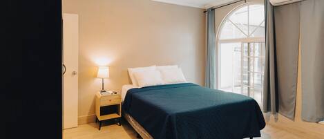 A bedroom with a bed, a nightstand with a lamp, a wooden floor, and a window with curtains.