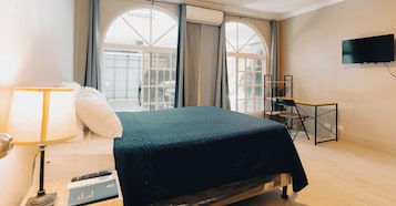 Habitación de hotel con cama, escritorio, silla, televisor y ventanas amplias.