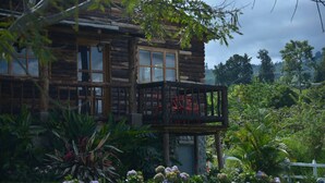 A rustic log cabin with a balcony, surrounded by lush greenery and flowering plants.