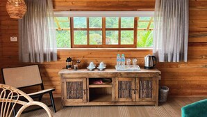 A cozy room with a wooden cabin-style interior, featuring a window with curtains, a wooden cabinet with kitchen items, a wicker chair, and a teal sofa.