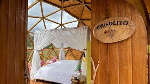 A wooden cabin bedroom with a bed, a canopy, a wooden sign with 'CRISOLITO' on it, and a view of greenery outside.