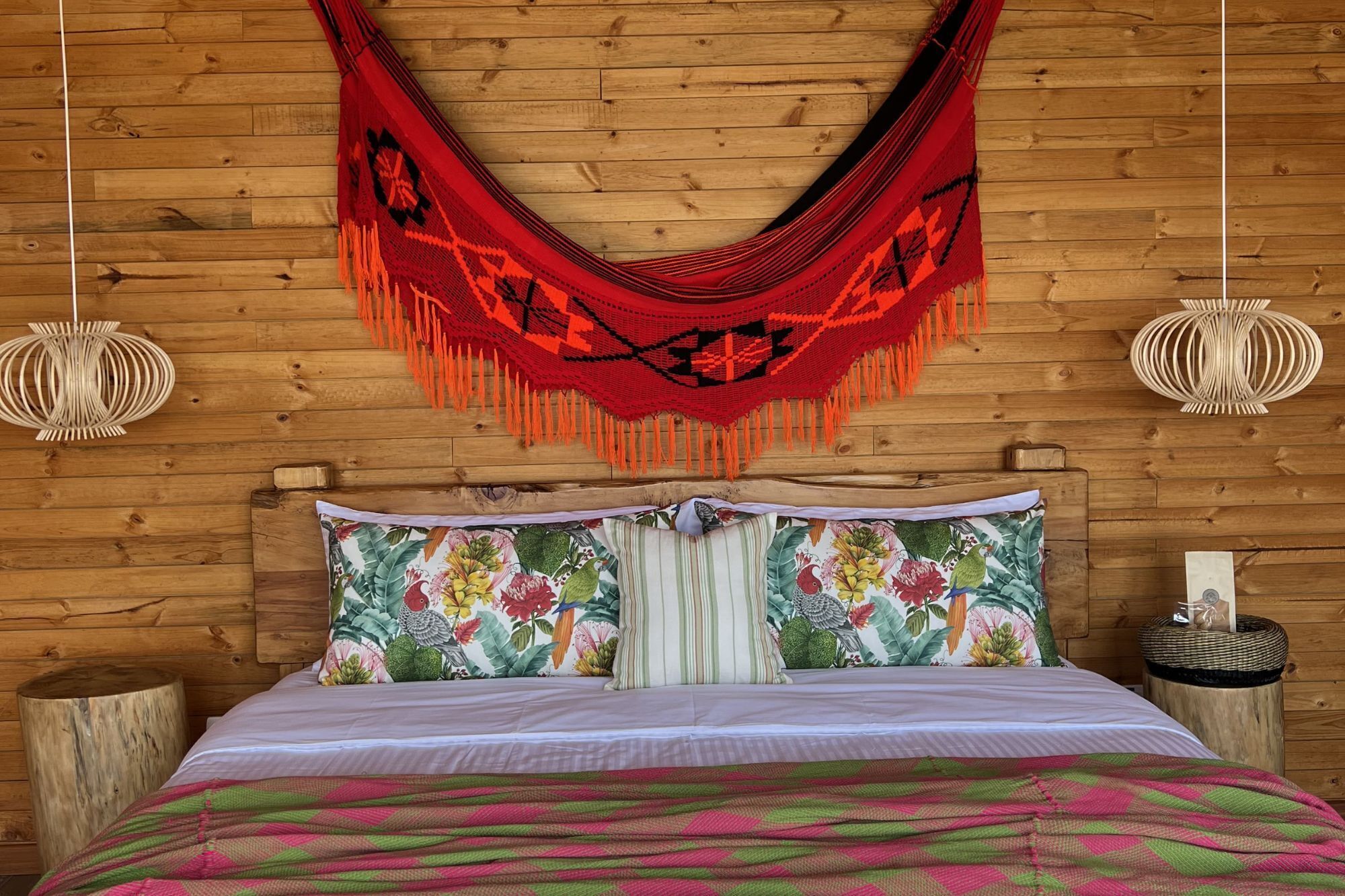 A bed with a floral pillow set, a red woven blanket, and a wooden headboard against a wooden wall.