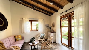 A cozy living room with a wooden ceiling, a pink sofa, wicker chairs, a small round table with a plant, and large windows with white curtains.